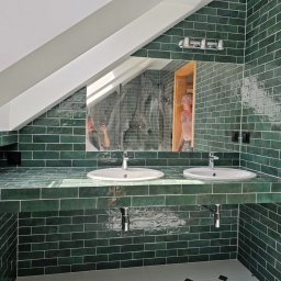FENIKS INVEST Sp. z o.o. - Łazienka na poddaszu wykończona ciemnozielonymi, błyszczącymi płytkami typu metro, z dwoma białymi umywalkami nablatowymi i dużym lustrem, w którym odbija się fragment prysznica i osoba.