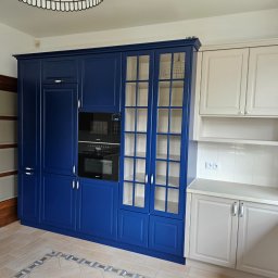 FENIKS INVEST Sp. z o.o. - Nowoczesna kuchnia z zabudową w kolorze granatowym i kremowym, z wbudowanymi piekarnikami Miele, oświetleniem sufitowym i podłogą z płytek ceramicznych z dekoracyjnym wzorem.