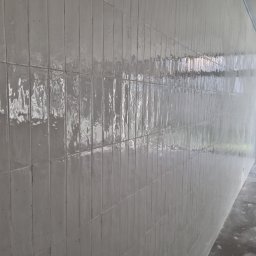 FENIKS INVEST Sp. z o.o. - Ściana wyłożona pionowymi, błyszczącymi, kremowymi płytkami o nieregularnej powierzchni, widoczne fugi, surowy beton na podłodze, w tle okno z widokiem na zieleń.