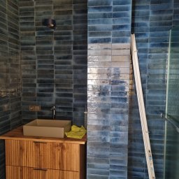 FENIKS INVEST Sp. z o.o. - Niebieska, błyszcząca glazura ułożona w cegiełkę na ścianach łazienki, z umywalką nablatową na drewnianej szafce i drabina oparta o ścianę obok kabiny prysznicowej.