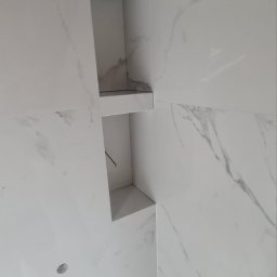 FENIKS INVEST Sp. z o.o. - Ściana wyłożona białymi płytkami imitującymi marmur z wnękami, w jednej z wnęk widoczny przewód elektryczny.