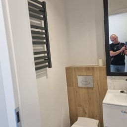 FENIKS INVEST Sp. z o.o. - Nowoczesna łazienka z wiszącą toaletą, grzejnikiem drabinkowym i umywalką z szafką, widoczne drewnopodobne płytki na ścianie za toaletą, odbicie fotografa w lustrze.
