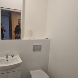 FENIKS INVEST Sp. z o.o. - Wykończona łazienka w minimalistycznym stylu, z białą umywalką na szafce, toaletą i lustrem, w którym odbija się osoba trzymająca telefon.