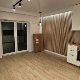 FENIKS INVEST Sp. z o.o. - Wnętrze po remoncie z drewnianą ścianą z lameli, podłogą imitującą drewno i białymi meblami kuchennymi. Duże okno balkonowe z widokiem na inne pomieszczenie. Na podłodze stoi nierozpakowany...