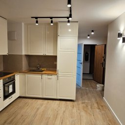 FENIKS INVEST Sp. z o.o. - Nowoczesna kuchnia z białymi szafkami, drewnianym blatem i panelami ściennymi, oświetlona reflektorami i kinkietami, z widocznym fragmentem przedpokoju z drzwiami wejściowymi.