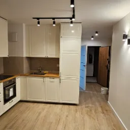 Nowoczesna kuchnia z białymi szafkami, drewnianym blatem i panelami ściennymi, oświetlona reflektorami i kinkietami, z widocznym fragmentem przedpokoju z drzwiami wejściowymi.