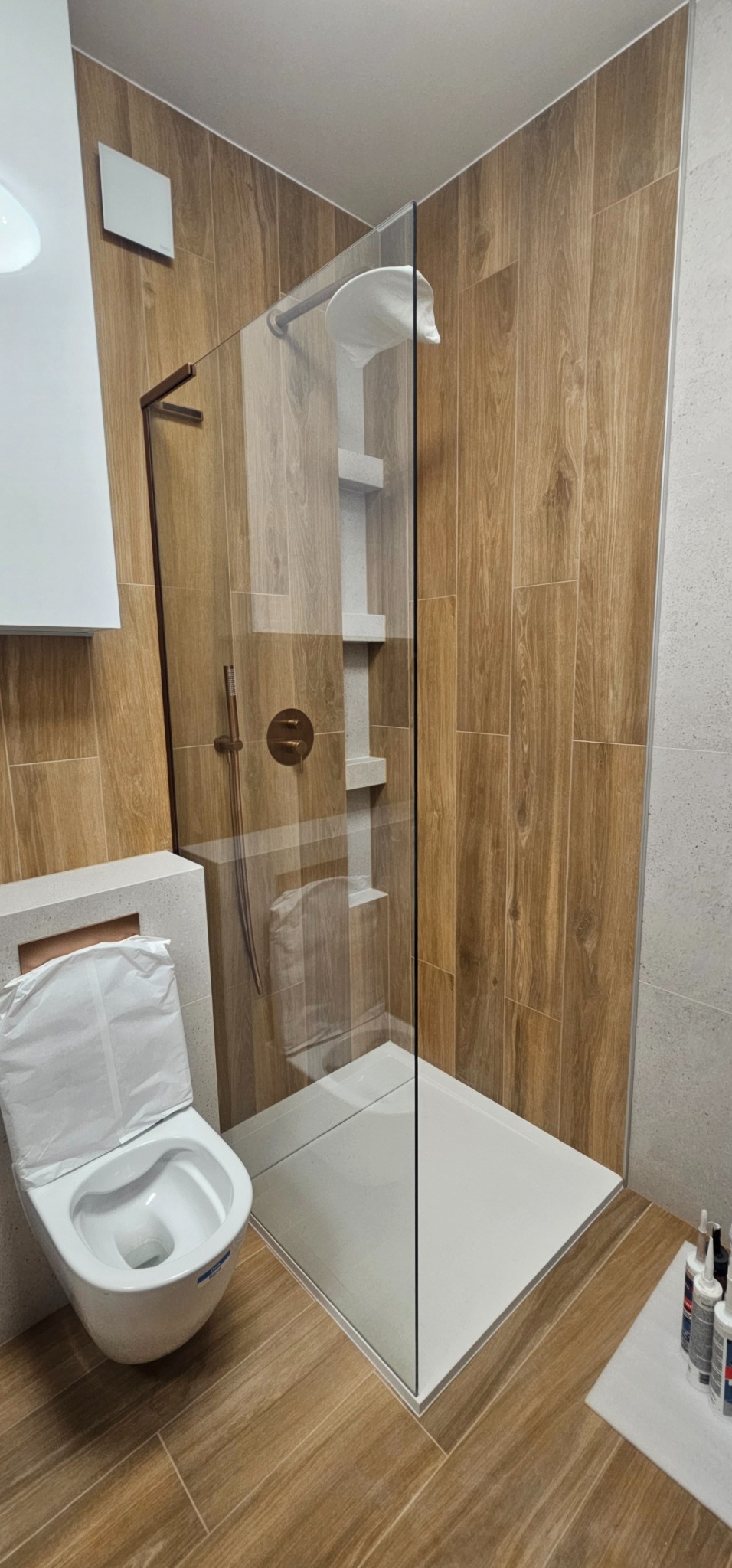 Nowoczesna łazienka z kabiną prysznicową typu walk-in, wykończona płytkami imitującymi drewno, z toaletą w trakcie montażu zabezpieczoną folią i brązową armaturą.