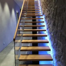 Nowoczesne schody z drewnianymi stopniami i szklaną balustradą, podświetlone niebieskim światłem, przylegające do ściany wyłożonej kamieniem.