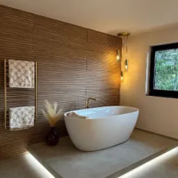 Nowoczesna łazienka z wolnostojącą białą wanną, złota bateria, drewniana ściana z grzejnikiem drabinkowym i ręcznikami JOOP!, podświetlana platforma pod wanną, wiszące lampy ze złotymi elementami...