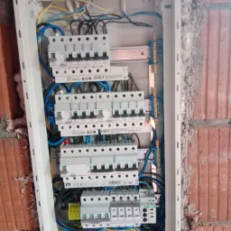 Otwarta skrzynka rozdzielcza z bezpiecznikami EATON i ETI, widoczne okablowanie w kolorach żółtym, niebieskim i czarnym, zamontowana na surowej ścianie z cegły.