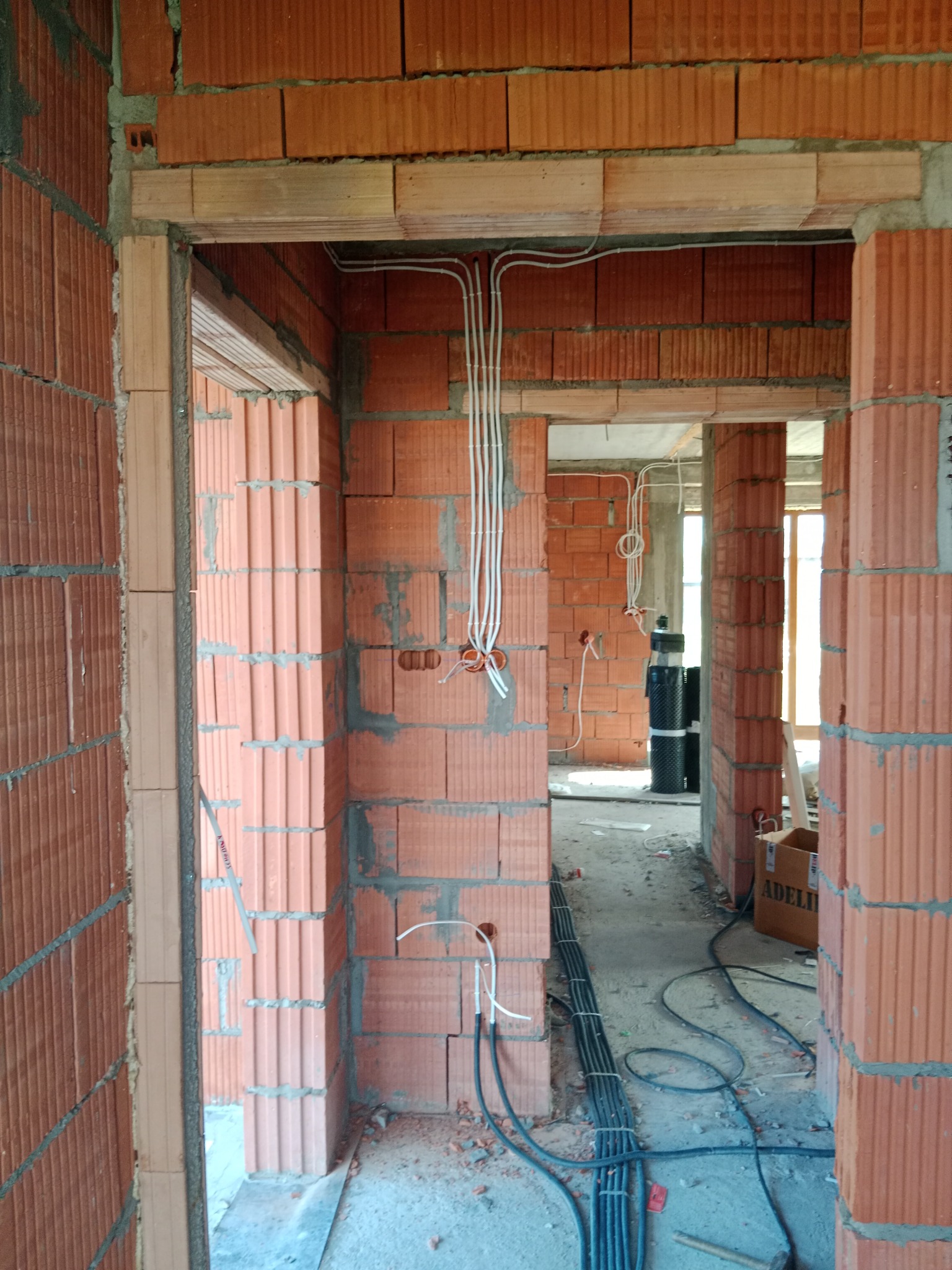 Wnętrze surowego budynku w trakcie prac elektrycznych: widoczne kable w białych peszlach prowadzone po ceglanych ścianach, przygotowane punkty pod gniazdka i włączniki, wiązka czarnych kabli...