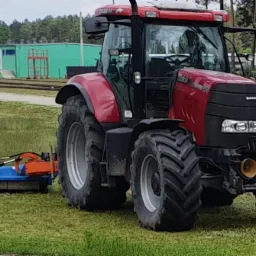 Czerwony traktor Case IH z podłączoną niebieską kosiarką bijakową Stark na trawiastym terenie, w tle tory kolejowe, budynek i pracownik w zielonej koszulce.