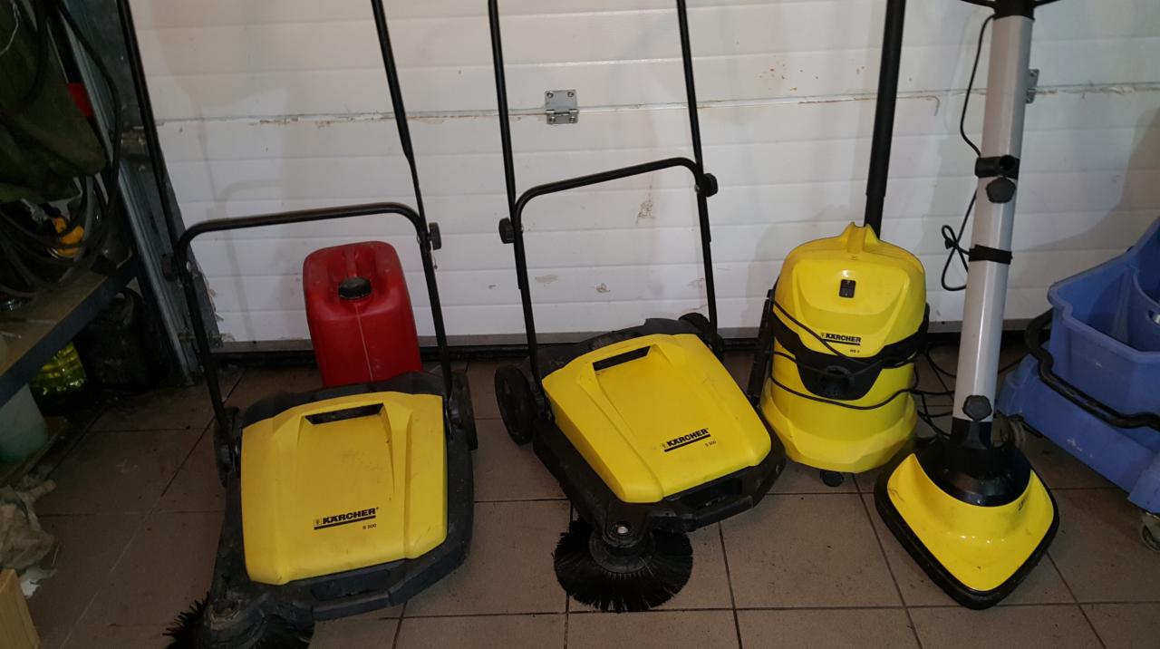 Żółte zamiatarki i odkurzacz przemysłowy Karcher stojące na brudnej podłodze z płytek w garażu, obok czerwony kanister i niebieski wózek do sprzątania.