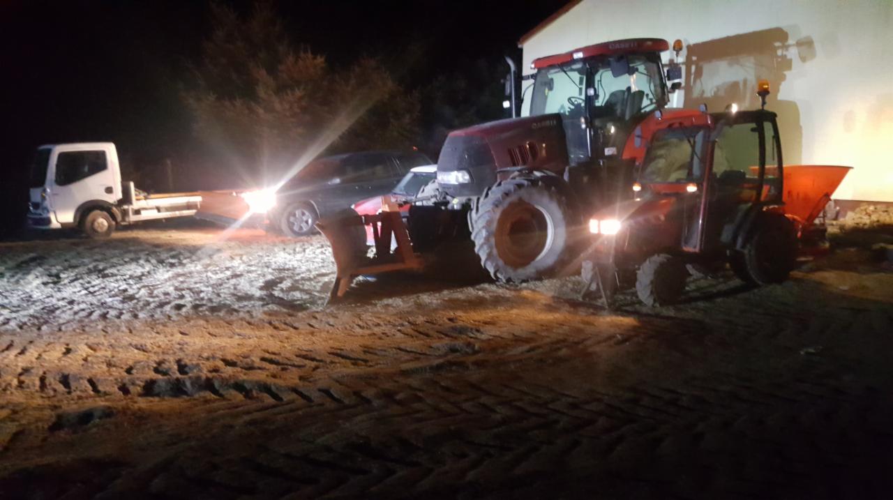 Dwa traktory, jeden z pługiem, drugi z rozsiewaczem, stoją na oświetlonym placu w nocy; w tle widoczny samochód i laweta.