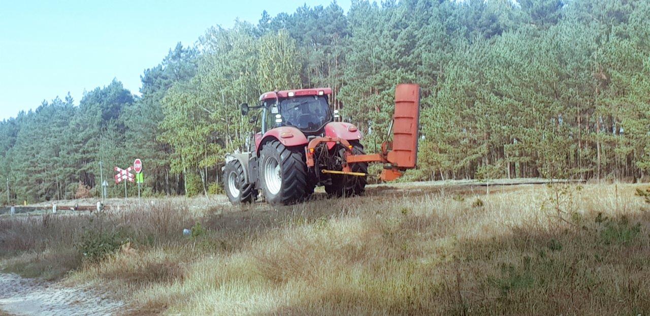 Czerwony traktor z kosiarką bijakową na polu z suchą trawą, w tle las iglasty i znak kolejowy.