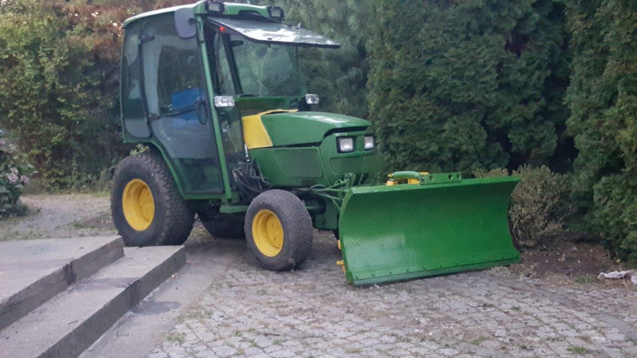 Zielony traktor z kabiną i żółtymi felgami, wyposażony w pług do odśnieżania, stojący na kostce brukowej przed zielonymi krzewami.