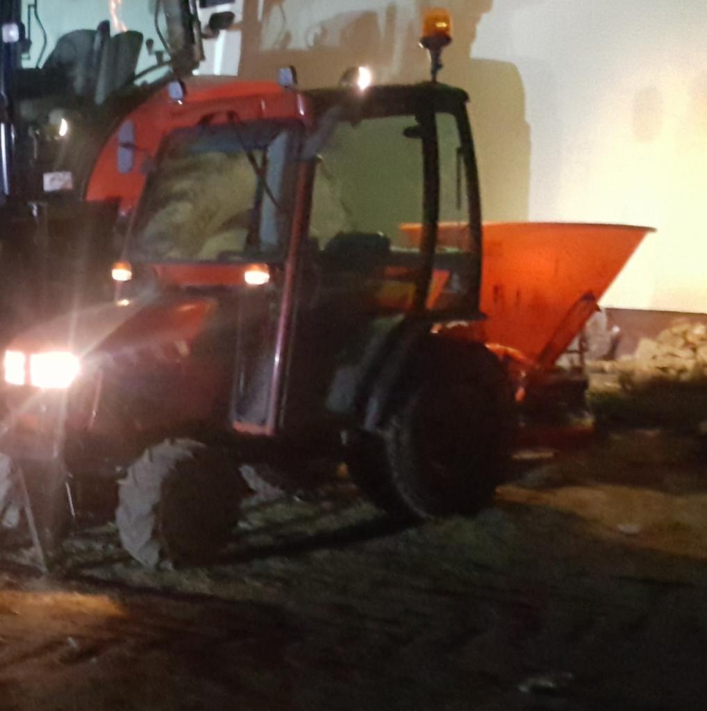 Pomarańczowy traktor z rozsiewaczem soli oświetlony reflektorami, stojący na ciemnym podłożu, prawdopodobnie w trakcie lub po posypywaniu nawierzchni.