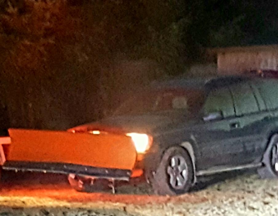 Ciemny SUV z pomarańczowym pługiem śnieżnym zamontowanym z przodu, oświetlony reflektorami, na tle ciemnego otoczenia.