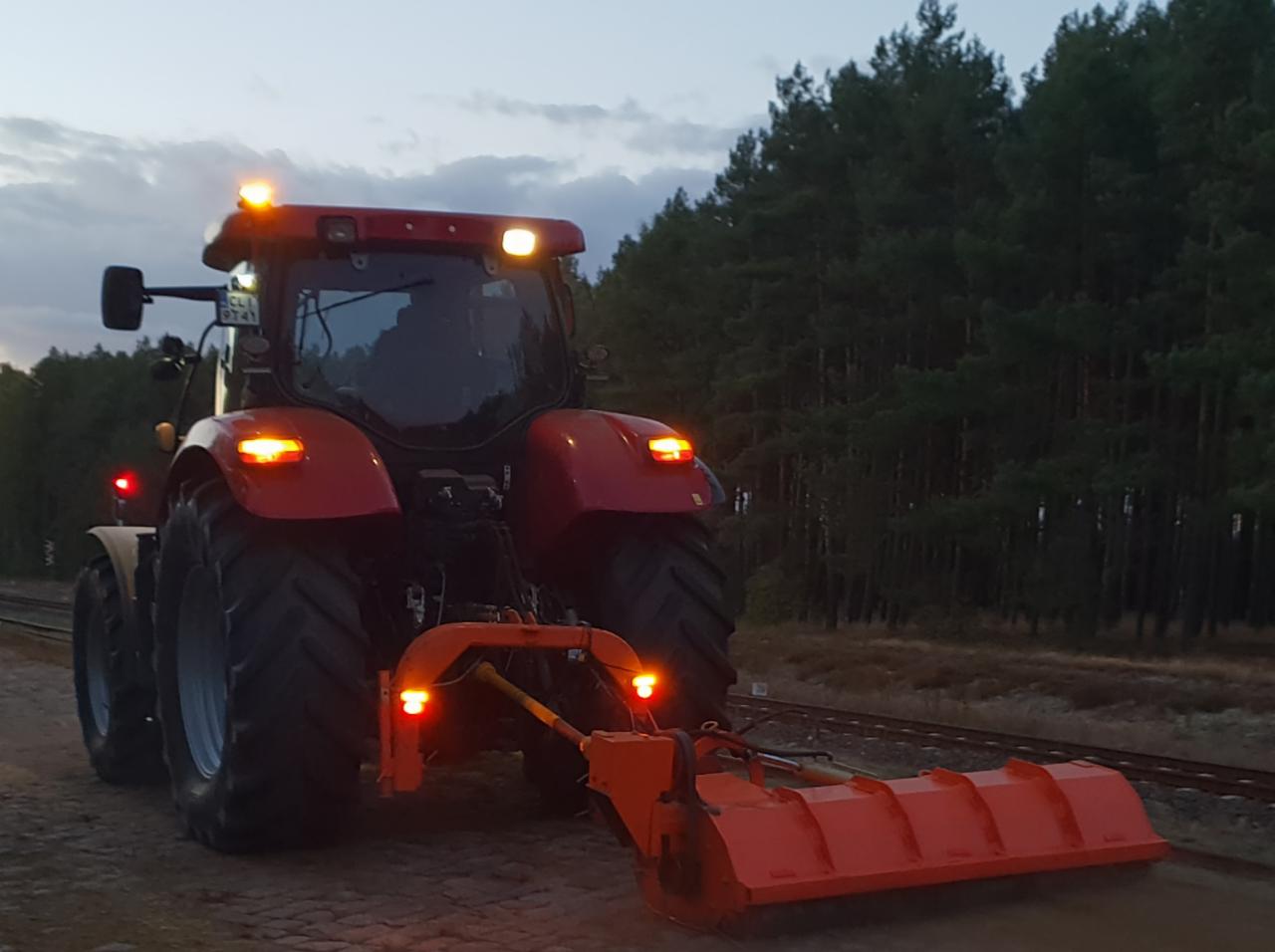 Czerwony traktor z zamontowanym pomarańczowym pługiem do odśnieżania, stojący na ubitej ziemi w pobliżu linii kolejowej, z lasem iglastym w tle i oświetlony zapalonymi światłami ostrzegawczymi.
