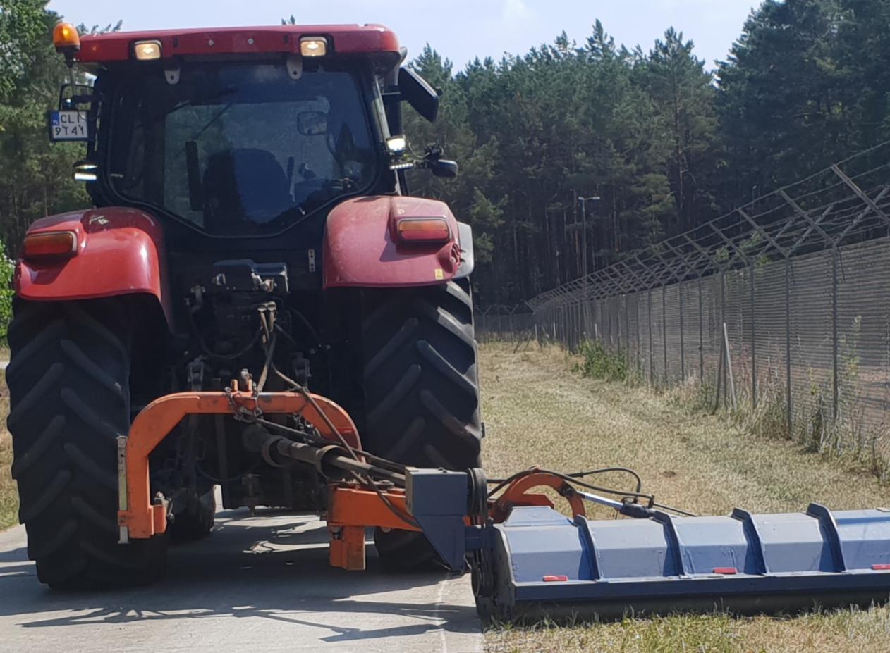 Czerwony traktor z kosiarką listwową na poboczu drogi, w tle siatka ogrodzeniowa i las.
