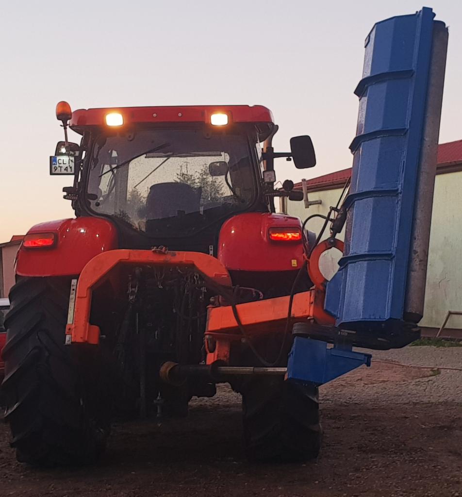 Czerwony traktor z niebieskim urządzeniem do pielęgnacji zieleni z tyłu, widok z tyłu, włączone światła, tablica rejestracyjna widoczna.