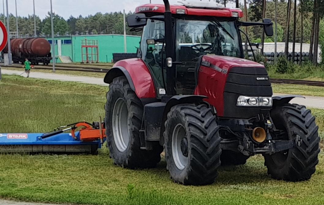 Czerwony traktor Case IH z podłączoną niebieską kosiarką bijakową Stark na trawiastym terenie, w tle tory kolejowe, budynek i pracownik w zielonej koszulce.