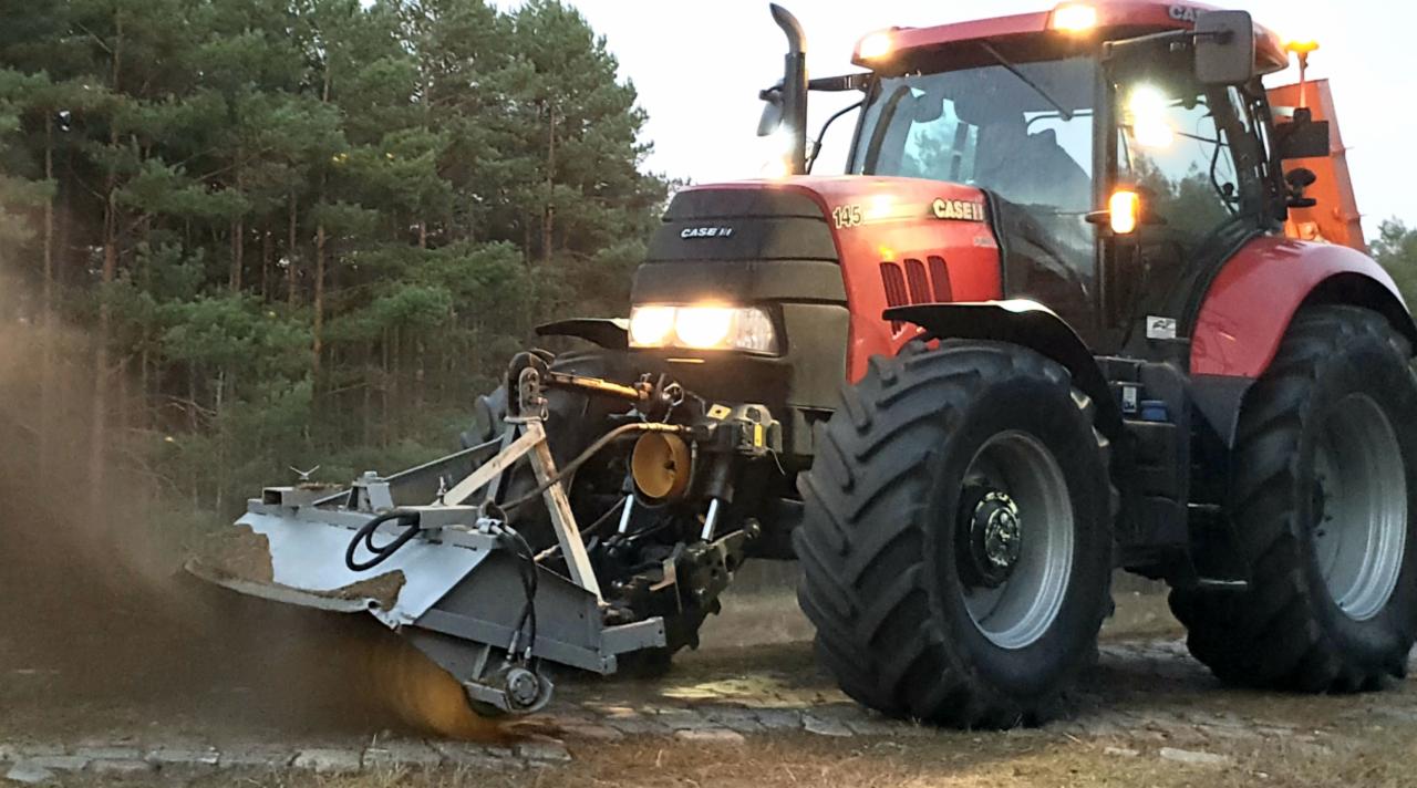 Czerwony traktor Case IH 145 z zamontowaną szczotką, usuwający piasek i zanieczyszczenia z leśnej drogi, podświetlony przez reflektory.