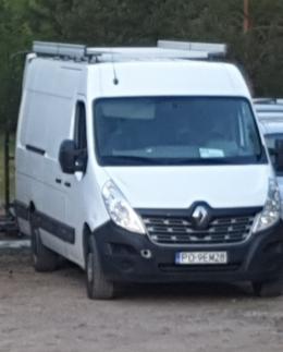 Biały samochód dostawczy Renault Master z bagażnikiem dachowym i widoczną tablicą rejestracyjną PO-9EM28, zaparkowany na szutrowym parkingu, częściowo zasłonięty przez inne pojazdy.