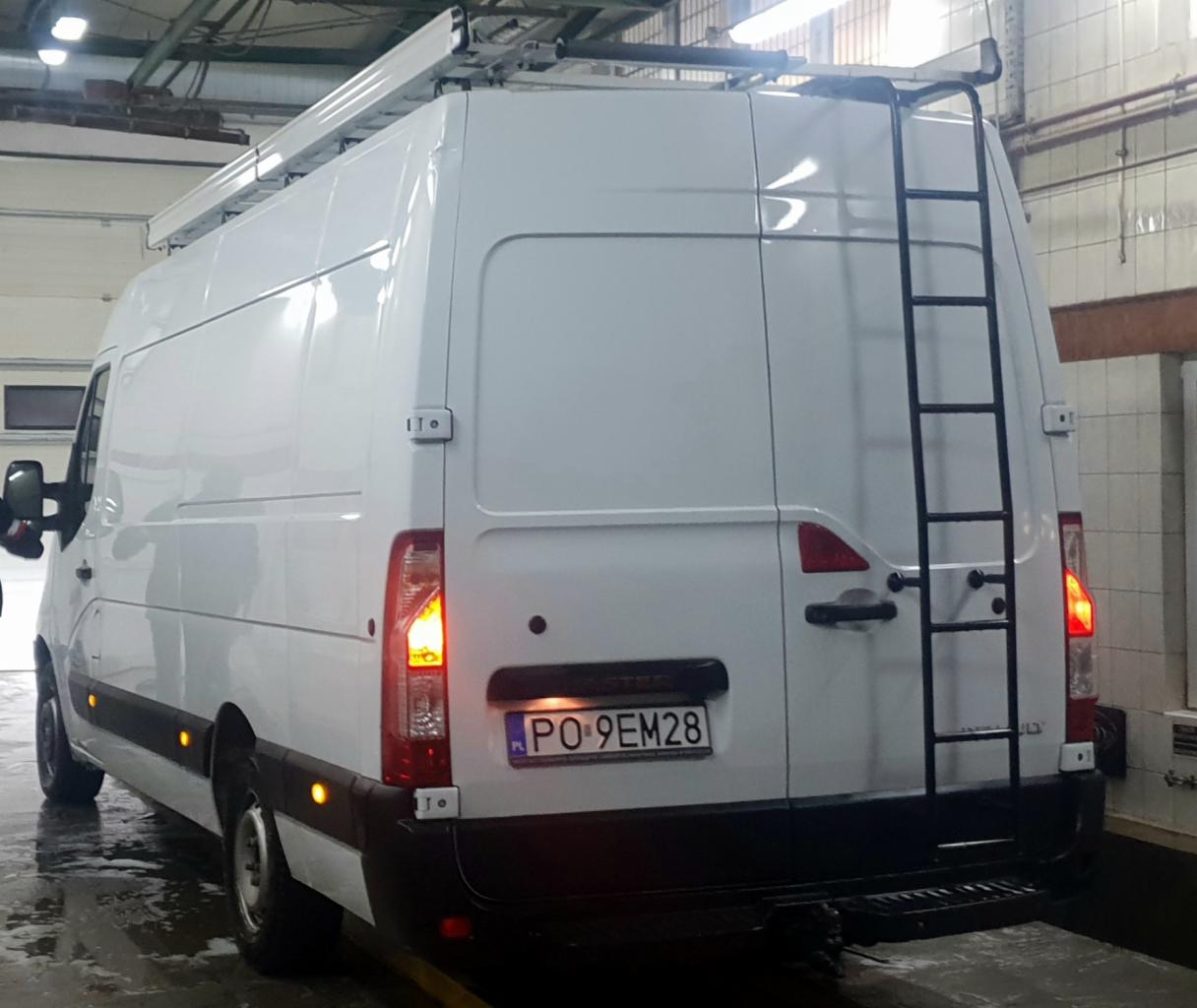 Biały samochód dostawczy z drabinką na tylnych drzwiach i relingami dachowymi, zaparkowany w hali z widoczną tablicą rejestracyjną.