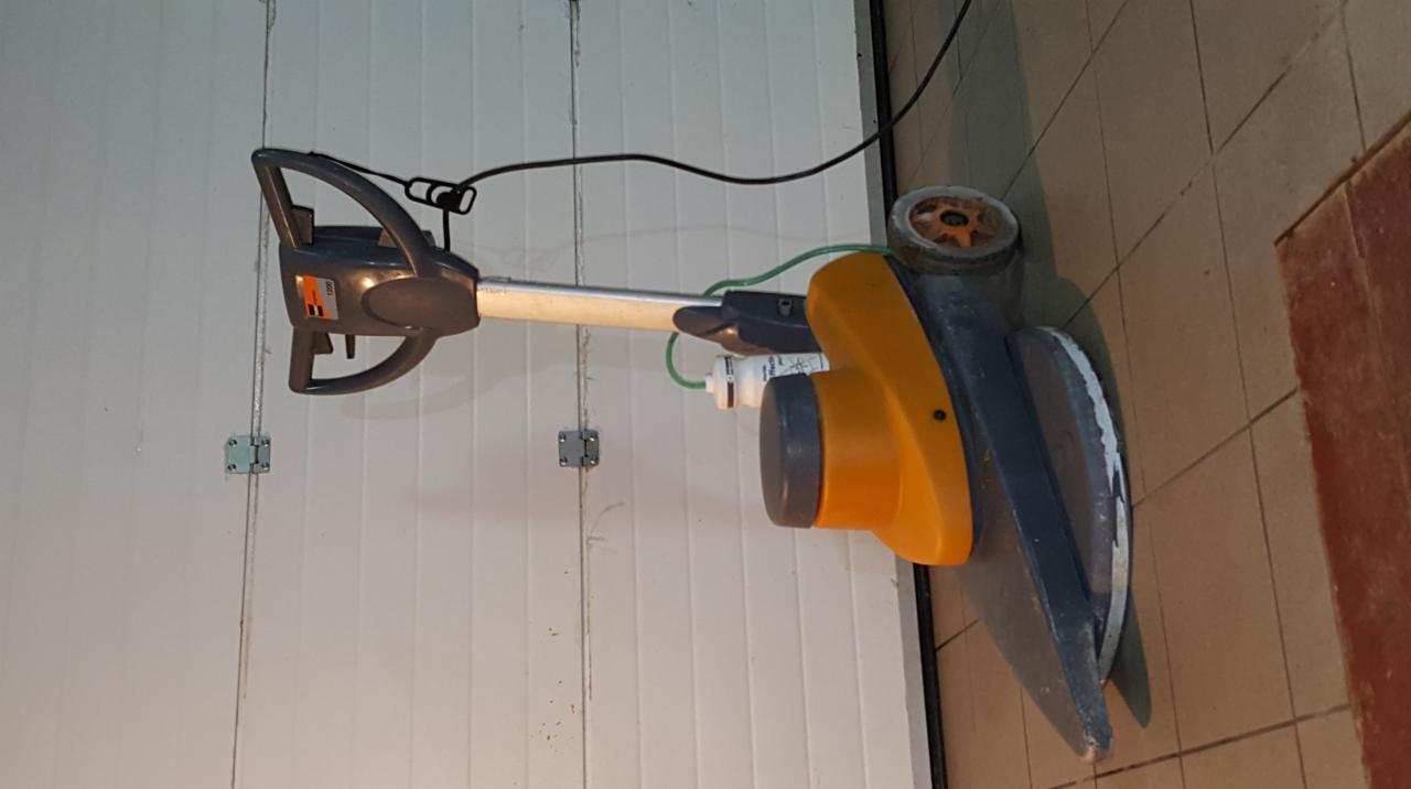 Elektryczna szorowarka do podłóg z żółtą obudową silnika i szarą szczotką, zaparkowana przy białej bramie garażowej i brązowych płytkach podłogowych, widoczne przewody zasilające.