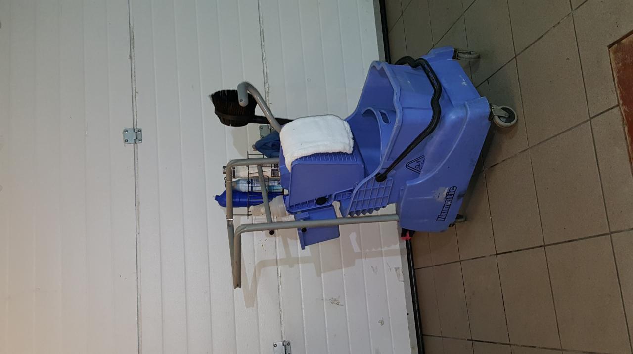 Niebieski wózek do sprzątania Numatic z mopem i szczotką, stojący na kafelkach przy białej ścianie.