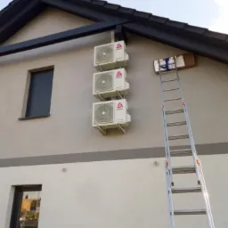 Trzy klimatyzatory marki CHIGO zamontowane jeden nad drugim na elewacji domu, obok rozstawiona aluminiowa drabina oparta o ścianę, z pudełkiem na górnym szczeblu.