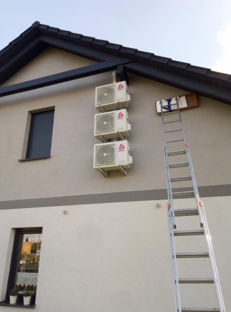 Trzy klimatyzatory marki CHIGO zamontowane jeden nad drugim na elewacji domu, obok rozstawiona aluminiowa drabina oparta o ścianę, z pudełkiem na górnym szczeblu.