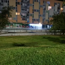 Nocny widok na blok mieszkalny z licznymi oknami, zieleń trawnika i drzewa na pierwszym planie, oświetlony pasaż handlowy w parterze budynku.