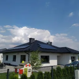 Biały dom jednorodzinny z czarnym dachem i panelami słonecznymi, widok zza zielonego żywopłotu, w tle błękitne niebo z obłokami.