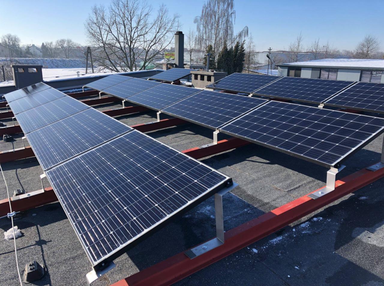 Panele fotowoltaiczne na płaskim dachu, zamontowane na czerwonej konstrukcji wsporczej, z widocznym śniegiem na dachu sąsiedniego budynku i drzewami w tle.