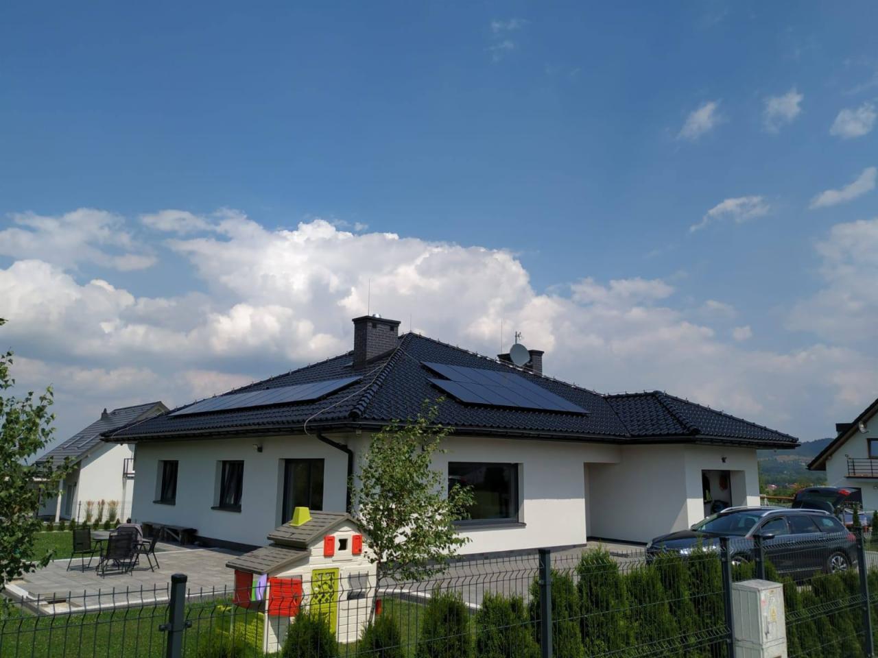 Biały dom jednorodzinny z czarnym dachem i panelami słonecznymi, widok zza zielonego żywopłotu, w tle błękitne niebo z obłokami.