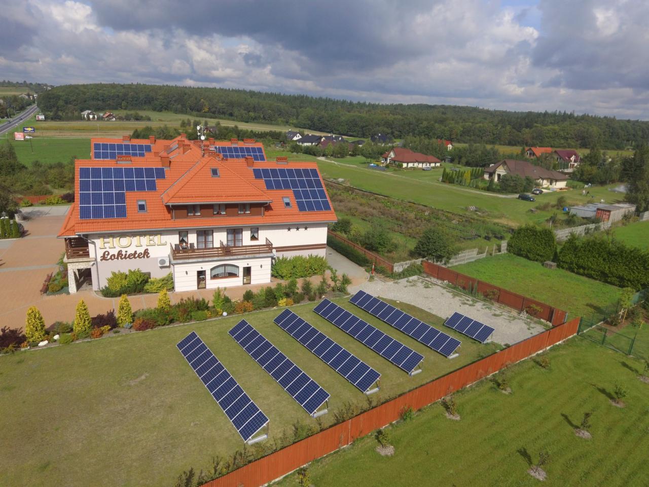 Hotel z panelami słonecznymi na dachu i rzędach paneli na trawniku, widok z góry na budynek z pomarańczowym dachem i okoliczne tereny zielone.