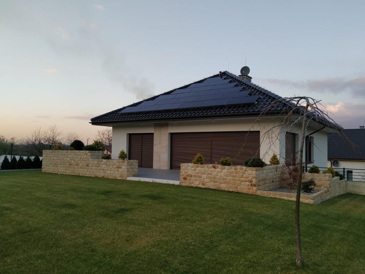 Nowoczesny dom jednorodzinny z panelami słonecznymi na dachu, widok z boku na zadbany trawnik i kamienne murki.