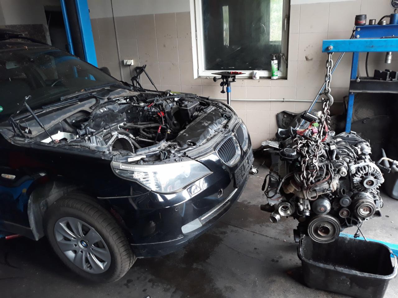 Czarny samochód BMW z otwartą maską, widoczny silnik zawieszony na łańcuchu obok pojazdu w warsztacie samochodowym.