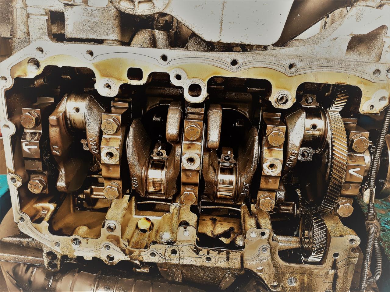 Olejem pokryty, otwarty silnik samochodowy z widocznymi wałami korbowymi, śrubami i przekładniami zębatymi, ukazujący wewnętrzne mechanizmy w trakcie naprawy lub konserwacji.