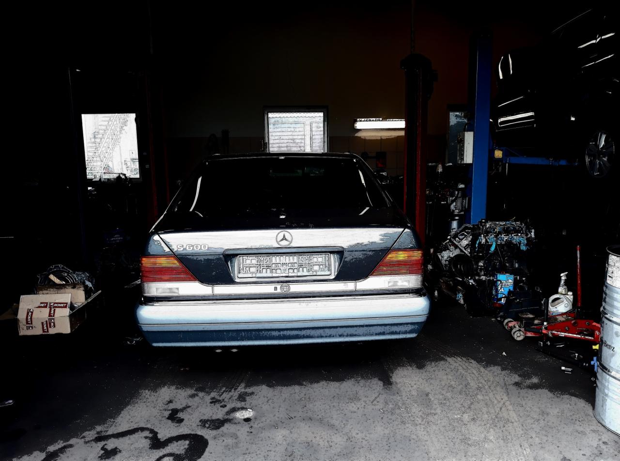 Tył ciemnego Mercedesa S600 w warsztacie samochodowym; widoczny silnik na wózku i podnośnik samochodowy w tle.