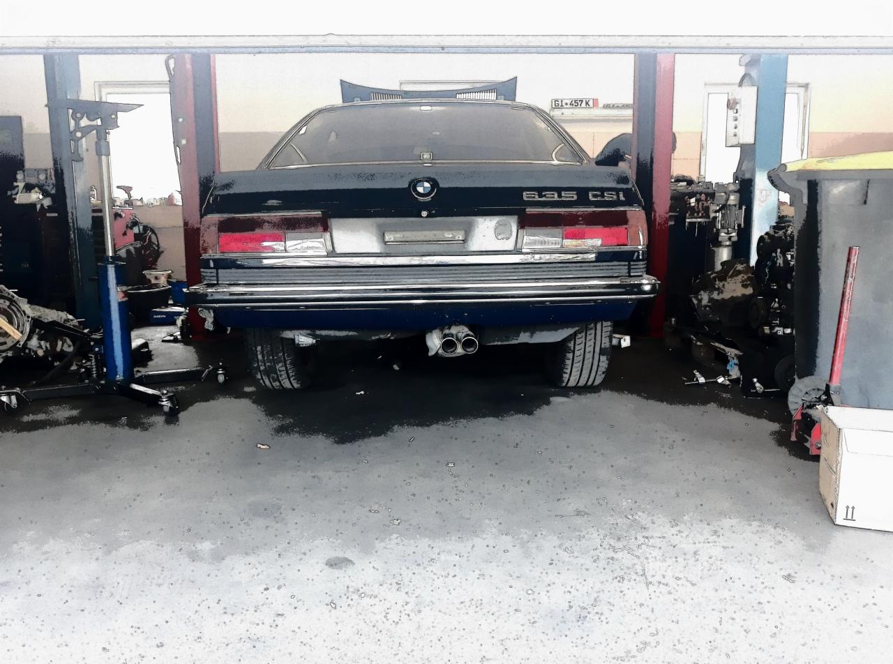 Tył granatowego BMW 635 CSi na podnośniku w warsztacie, widoczne podwójne rury wydechowe, tablica rejestracyjna GI 457K, otwarty bagażnik, na podłodze plamy oleju i części samochodowe.