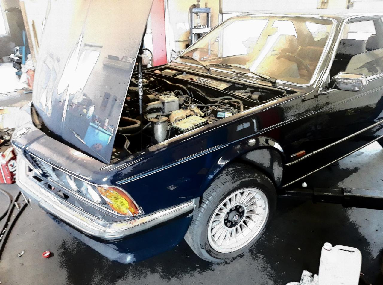 Niebieski samochód z otwartą maską w warsztacie, widoczny silnik i przednie koło z felgą w stylu retro.