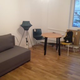 Remonty Wykonczeinie - Pusta biała ściana w pokoju po malowaniu, widoczna szara sofa, stół z czarnymi nogami i krzesła, lampa na trójnogu i drewniana podłoga.