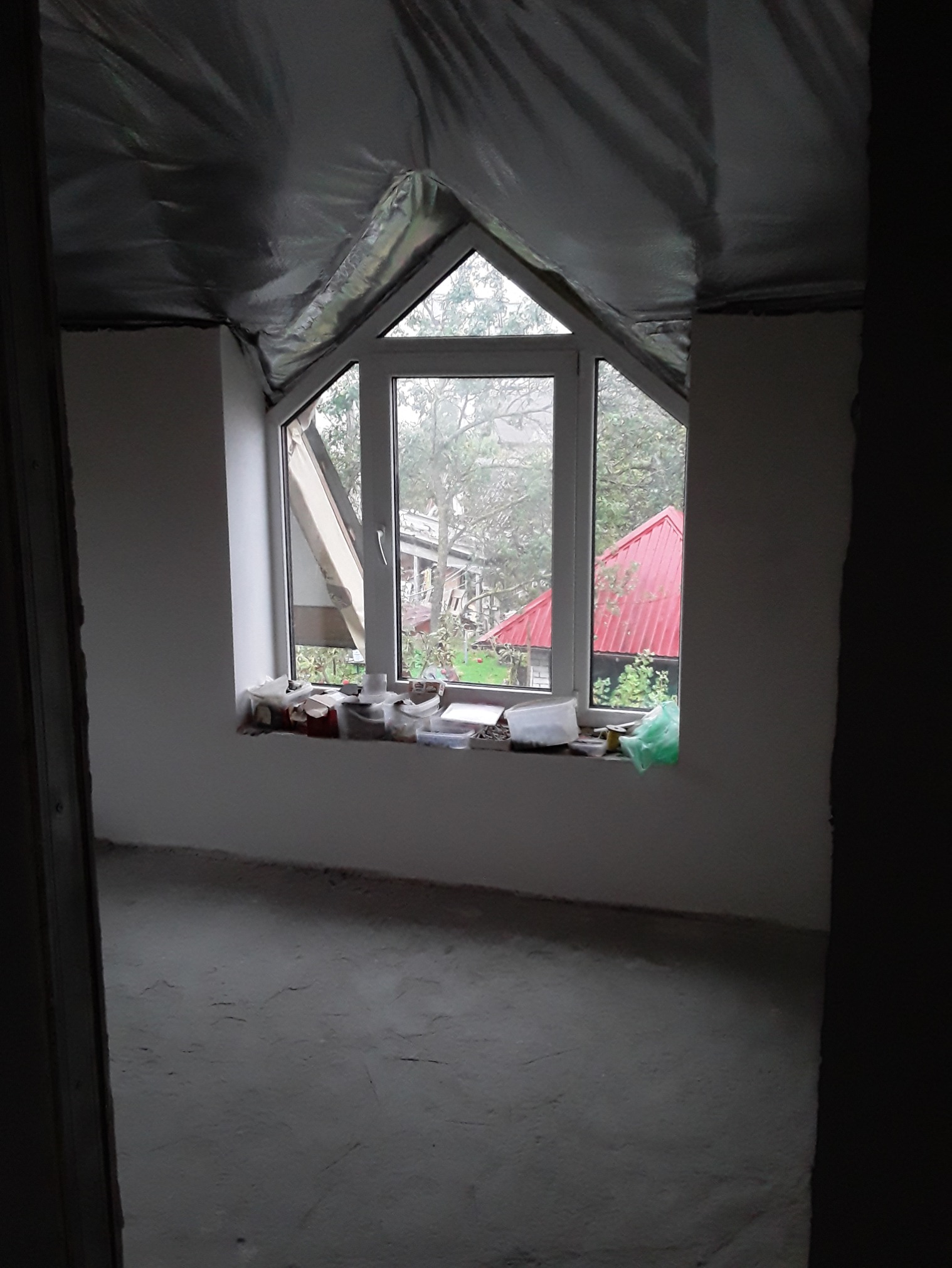Pokój na poddaszu w trakcie remontu: surowy beton na podłodze, ściany częściowo wygładzone, okno dachowe z widokiem na ogród, folia izolacyjna na skośnym suficie.