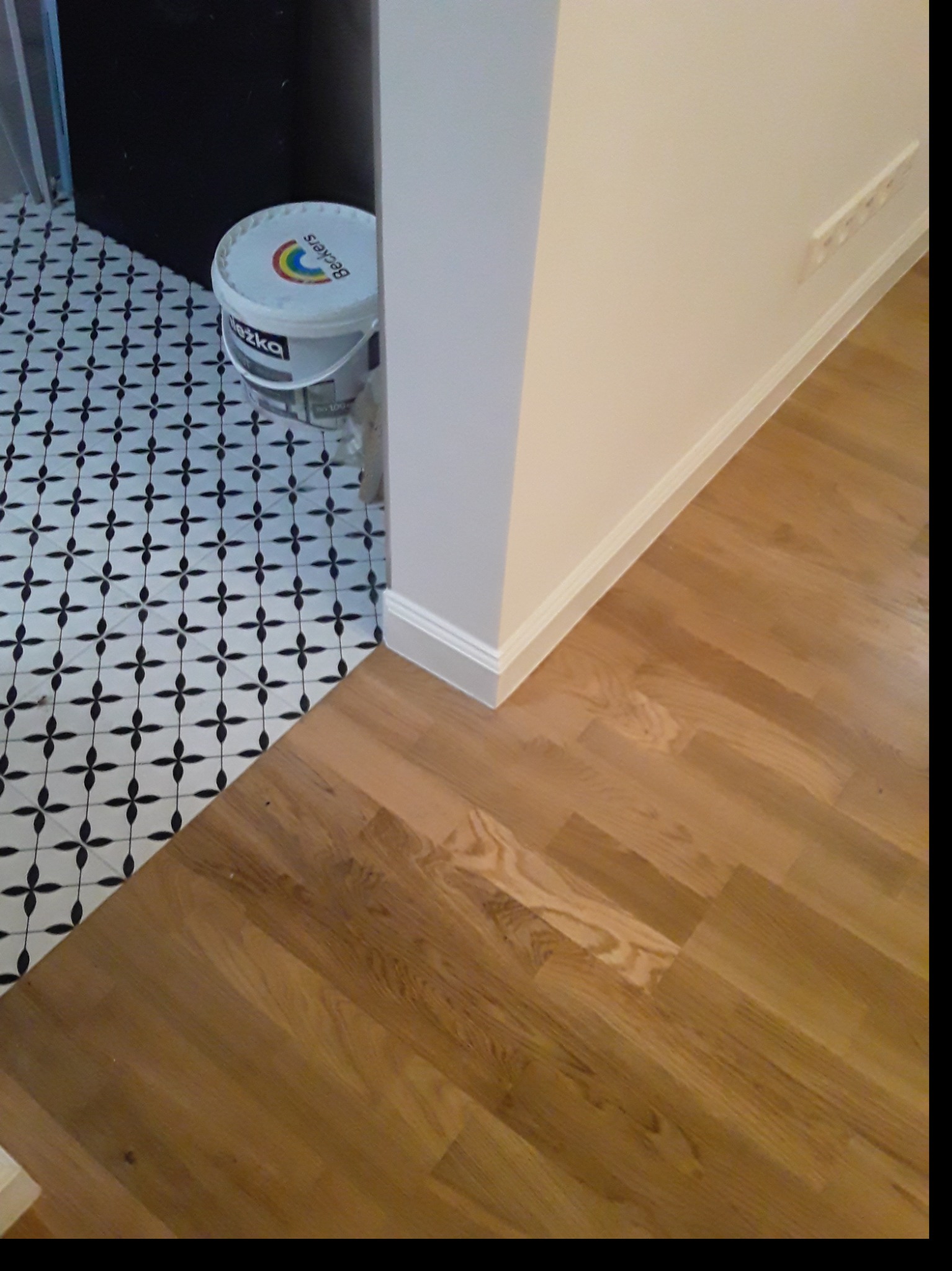 Fragment wykończonego wnętrza z kontrastującym połączeniem podłóg: drewniany parkiet i czarno-biała mozaika, przy ścianie stoi wiadro z farbą marki Beckers.