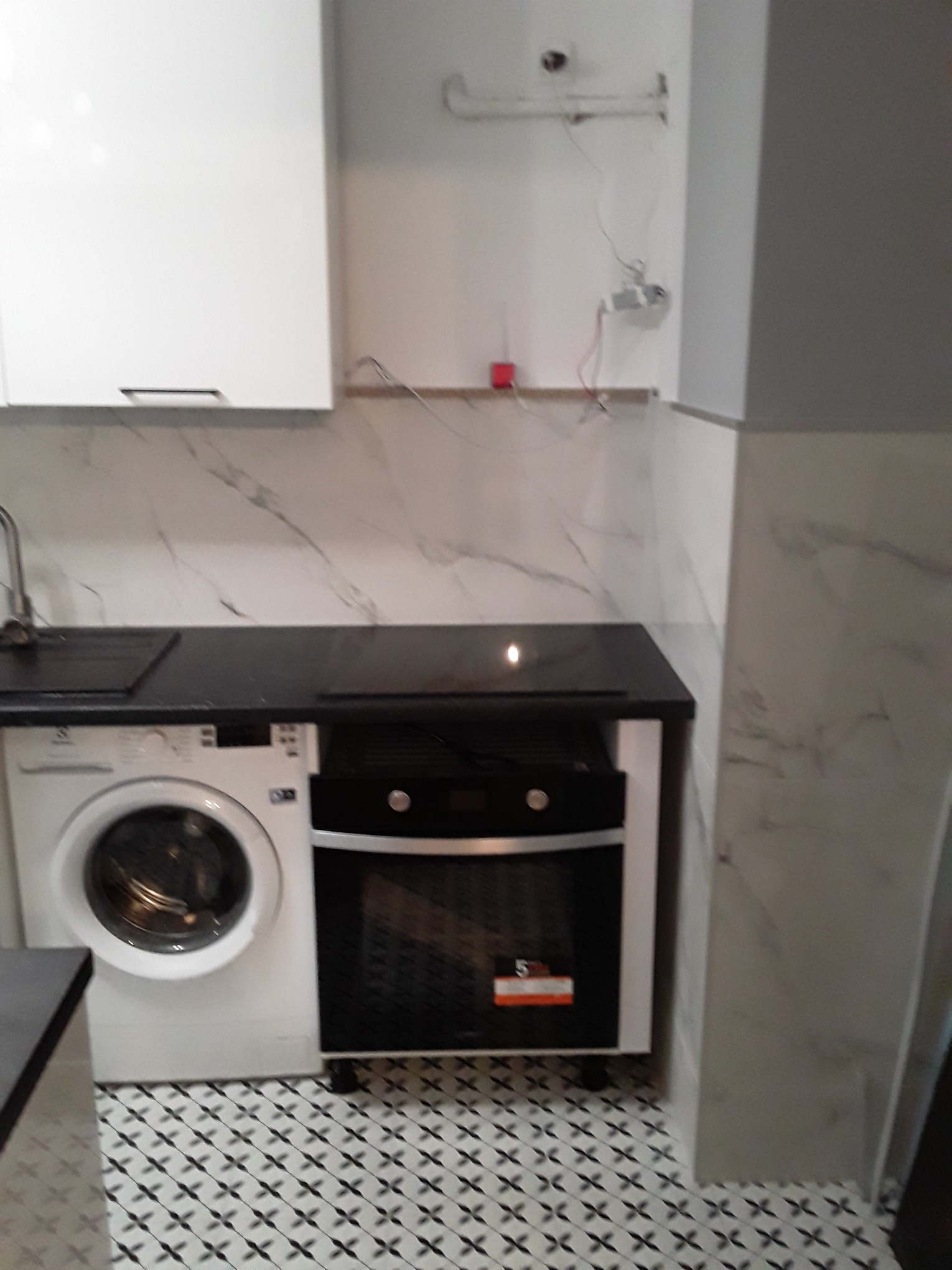 Kompaktowa zabudowa kuchenna z pralką obok piekarnika, czarny blat i marmurowe płytki na ścianie.