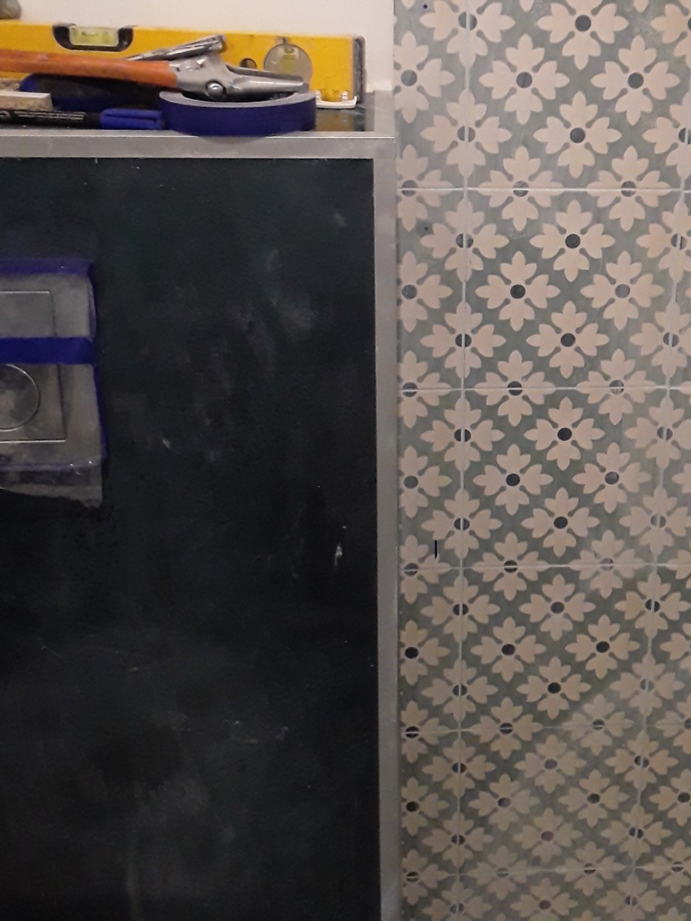 Fragment łazienki w trakcie prac: widoczne płytki ścienne z wzorem kwiatowym, czarna zabudowa stelaża WC, poziomica i narzędzia na górze stelaża.