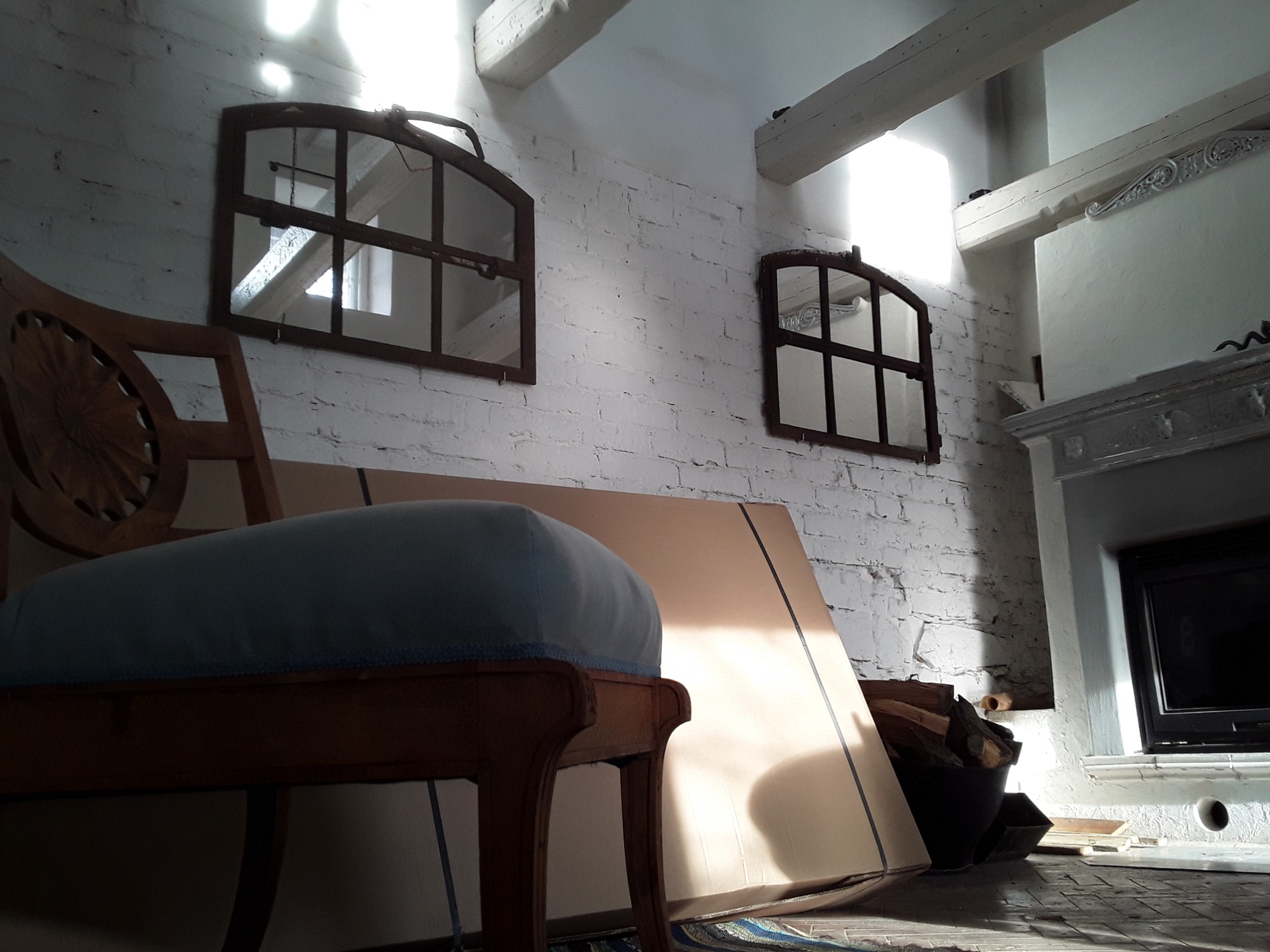Wnętrze w trakcie remontu: drewniane okna-lustra na białej ceglanej ścianie, karton gips oparty o ścianę, fragment krzesła z niebieskim siedziskiem, kominek z czarnym wkładem i drewno w wiadrze.